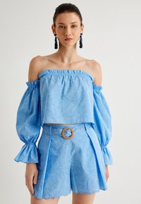 Top corto azul claro con volantes en los hombros, mangas voluminosas, tejido texturizado y patrones circulares blancos. Combina con pantalones cortos y cinturón a juego.