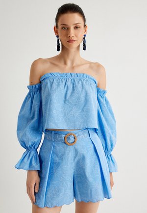 Lichtblauw off-shoulder crop top met volumineuze mouwen, textuurstof en witte cirkelpatronen. Gecombineerd met bijpassende shorts en riem.