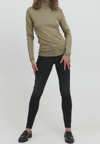 Olivgrön högkragetröja med långa ärmar, kombinerad med svarta slim-fit jeans och svarta loafers, mot en enfärgad bakgrund.