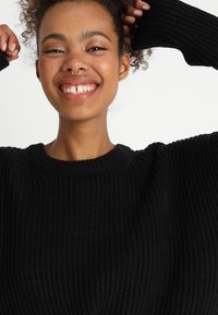 Schwarzer gerippter Pullover aus Strick mit rundem Halsausschnitt und weiten Ärmeln, der einen strukturierten Stoff zeigt. Es gibt keine zusätzlichen Muster oder Hardware.
