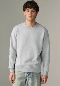 Sudadera gris claro de mangas largas, escote redondo, con puños y dobladillo acanalados, combinada con jeans azul claro. Textura suave, ajuste casual.