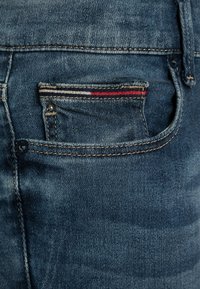 Vaqueros de mezclilla con un lavado azul oscuro, que presentan un pequeño bolsillo trasero con acentos de rayas en rojo, blanco y azul y costuras contrastantes.