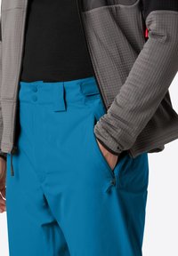 Pantaloni softshell blu con una superficie liscia, dotati di due chiusure a bottone e una tasca laterale con zip, abbinati a una giacca grigia testurizzata.