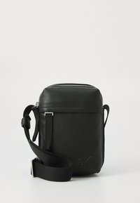 Black leather crossbody bag with textured surface, silver zipper, adjustable strap, and embossed logo on the front. Compact rectangular shape.