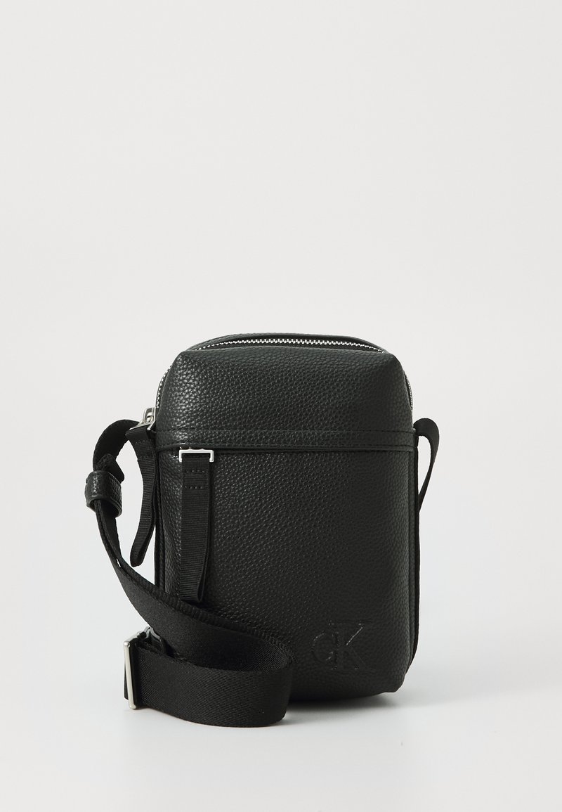 Black leather crossbody bag with textured surface, silver zipper, adjustable strap, and embossed logo on the front. Compact rectangular shape.