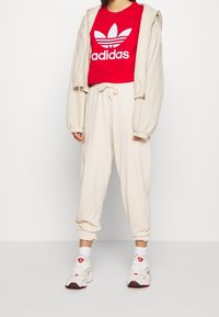 Červené zkrácené tričko s bílým logem Adidas, krémová mikina s kapucí a volné krémové tepláky, doplněné bílými a červenými sportovními botami.