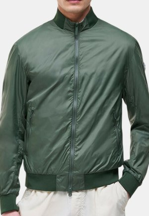Uomo che indossa una giacca bomber leggera verde oliva con zip, collo, polsini e fascia alla vita a coste, abbinata a pantaloni chiari.