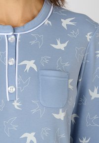 Camisetta azzurra chiara con motivi di uccelli bianchi, scollatura rotonda e taschino frontale. Presenta un sottile placket e una rifinitura a contrasto.