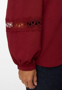 Bordeaux blouse met lange, opbollende mouwen. Heeft een delicate kantafwerking bij de manchetten. Zachte, textuurstof met elastische mouwuiteinden.