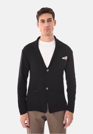 Uomo che indossa un blazer nero abbottonato con fazzoletto da taschino fantasia, maglietta bianca a girocollo e pantaloni marroni su sfondo neutro.