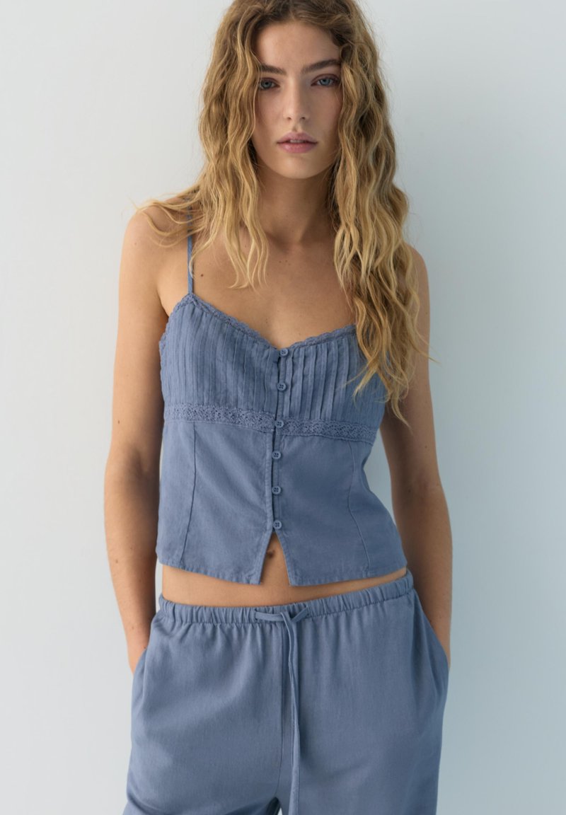 Jeune femme aux longs cheveux ondulés portant un haut bleu sans manches avec des boutons et un pantalon assorti à cordon, debout devant un fond clair uni.