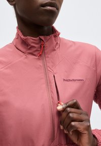 Άτομο που φοράει ροζ μπουφάν Peak Performance, τραβάει προς τα πάνω το φερμουάρ σε κάθετη τσέπη στήθους με κόκκινο κορδόνι.