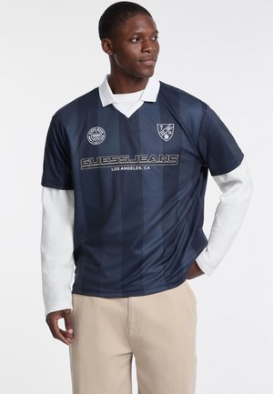 Marineblau gestreiftes Polohemd mit weißem Kragen, verziert mit dem GUESS JEANS Logo und Aufnähern. Lange weiße Ärmel darunter.