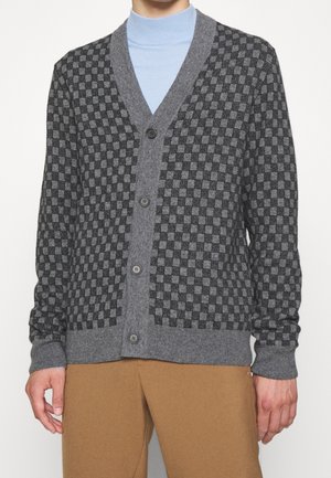 Homme portant un cardigan gris à carreaux boutonné par-dessus un col roulé bleu clair et un pantalon marron, debout devant un fond uni.