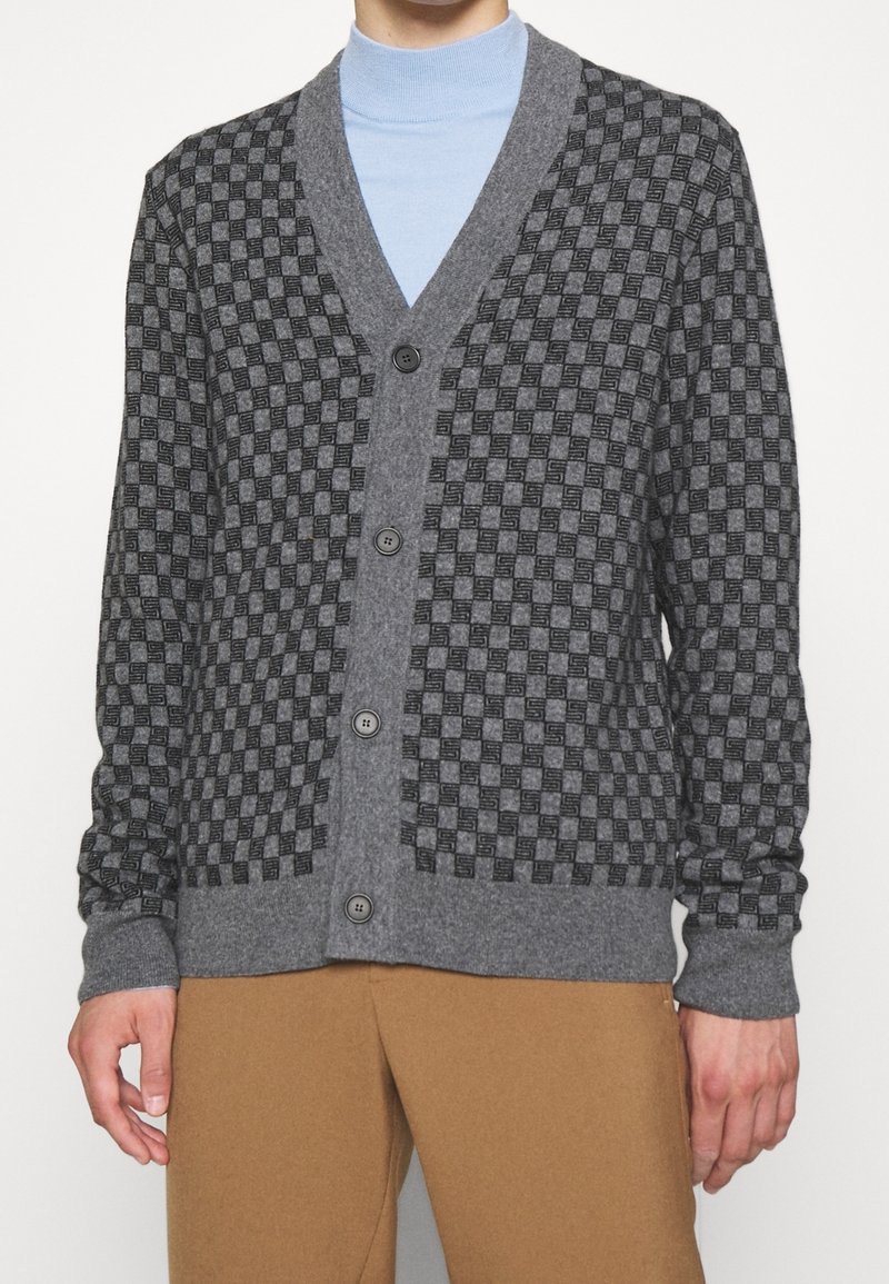 Homme portant un cardigan gris à carreaux boutonné par-dessus un col roulé bleu clair et un pantalon marron, debout devant un fond uni.