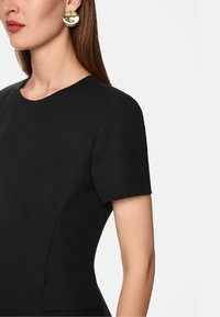 Schwarzes, kurzärmliges Kleid mit Rundhalsausschnitt, das eine taillierte Form und glatte Textur aufweist. Goldene, runde Ohrringe setzen einen Kontrast und setzen Akzente.