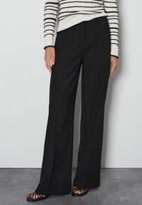 Pantalons larges noirs à la coupe ajustée, avec une texture lisse et des lignes épurées, associés à des chaussures noires à lanières et un haut rayé.