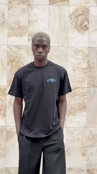 Camiseta negra con un logo turquesa, de manga corta y corte relajado, combinada con pantalones oscuros y holgados, contra un fondo texturizado.