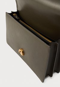 Χειροποίητη τσάντα ώμου από δέρμα σε ελαιώδες πράσινο χρώμα με κούμπωμα χρυσού κουμπιού και τσέπη με φερμουάρ. Ομαλή υφή, ορθογώνιο σχήμα, καθαρές γραμμές.