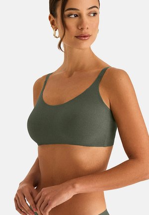Vrouw die een geribbelde olijfgroene bralette met dunne bandjes draagt, gouden oorringen en haar handen losjes gevouwen ter hoogte van de taille.