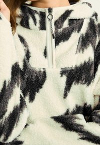 Fleecepullover i cremefarve med sort zigzagmønster. Har kvart-lynlås foran, høj krave og blødt, struktureret stof.