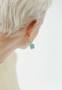 Pendiente de aro dorado con un charm rectangular de turquesa que presenta una letra "H" en relieve. El fondo es sencillo, lo que enfatiza los detalles del pendiente.