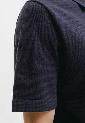 Gros plan d'une personne portant une chemise texturée bleu marine foncé à manches courtes avec un col plié, montrant la partie supérieure du bras et la zone de l'épaule.