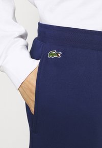 Personne portant un pantalon bleu marine avec un logo vert de crocodile sur la poche, main dans la poche et chemise blanche à manches longues visible.