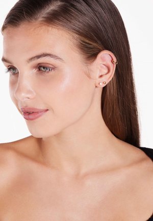 Gouden hoepeloorbel en twee studs met heldere edelstenen. Het model heeft lang, stijl haar en blote schouders tegen een witte achtergrond.