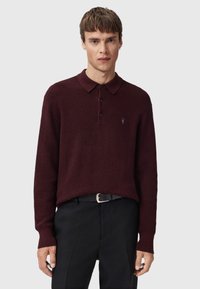 Burgundy knit pullover with a collar, three button placket, and small embroidered logo. Worn over black trousers and a belt.