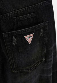 Schwarzer Denim mit einer Gesäßtasche, die einen dreieckigen rot-weißen Guess-Logo-Patch zeigt. Der Stoff weist eine leicht abgenutzte Textur auf.