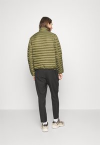 Olijfgroene pufferjack met horizontale quiltstiksel, hoge kraag en een ontspannen pasvorm, gecombineerd met lichtgrijze broeken en beige sneakers.