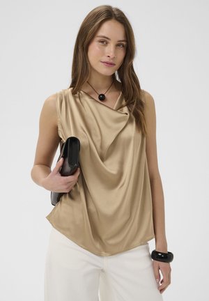 Nő bézs drapírozott, ujjatlan felsőben, fehér nadrágban, fekete karkötővel és nyaklánccal, fekete borítéktáskával egy sima háttér előtt.