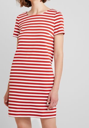 Kobieta w sukience z krótkim rękawem sięgającej kolan, w poziome czerwono-białe paski, stojąca na jednolitym jasnoszarym tle.