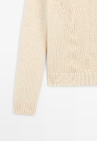 Pull en maille crème avec un ourlet et des poignets côtelés, présentant un motif texturé. Le matériau semble doux et chaud, adapté aux temps frais.