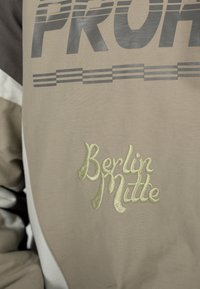 Veste beige et marron avec les mots "Berlin Mitte" brodés et un grand "PRUH" imprimé au-dessus avec des lignes horizontales.