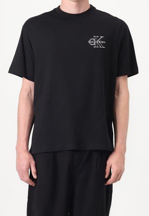 Mann trägt ein schwarzes kurzärmliges T-Shirt mit kleinem weißem Calvin Klein Logo auf der oberen linken Brust, kombiniert mit schwarzen Hosen.