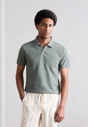 Άνδρας που στέκεται δίπλα σε ένα απλό τοίχο, φοράει γκρι-πράσινο πόλο με κοντό μανίκι και ανοιχτό μπεζ ριγέ παντελόνι με κορδόνι, με τα χέρια στις τσέπες.