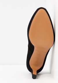 Czarny zamszowy but na wysokim obcasie w kształcie migdała pokazany od strony podeszwy, z gładką beżową podeszwą i małym czarnym czubkiem obcasa.