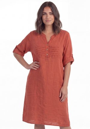 Vrouw met schouderlang bruin haar, gekleed in een roestkleurige, geknoopte, knielange jurk met opgerolde mouwen, staand met één hand aan de heup.