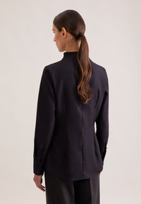 Blusa a maniche lunghe di colore blu navy scuro con collo alto, texture liscia e polsini con chiusura. Presenta una cucitura posteriore e una silhouette aderente.