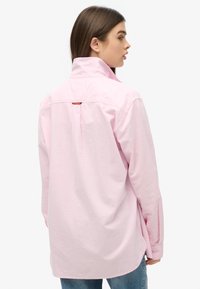 Jasnoróżowa koszula z długim rękawem, z zapiętą kołnierzykiem, pojedynczym plisowaniem z tyłu oraz małą metką markową w okolicy szyi; zestawiona z niebieskimi dżinsami.