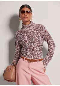 Langärmliger, Stehkragenpullover in Pink mit einem kunstvollen Paisley-Muster in Burgunderrot und Creme. Kombiniert mit hoch taillierten pinken Hosen und einem braunen Gürtel.