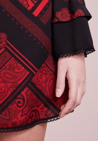 Czarna sukienka z czerwonymi wzorami paisley i geometrycznymi. Rozkloszowane rękawy i koronkowe wykończenie u dołu. Miękka tkanina o gładkiej fakturze.