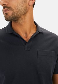 Dunkelblaues Poloshirt aus getexturem Stoff mit kragenloser Gestaltung und einer einzelnen Brusttasche. Stichdetails am Kragen.