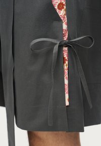 Κοντινό πλάνο ενός σκουρόχρωμου γκρίζου ενδύματος δεμένου με φιόγκο πάνω σε ένα υφασμάτινο με μοτίβο λουλουδιών σε ροζ και κόκκινους τόνους, που καλύπτει εν μέρει το πόδι ενός ατόμου.