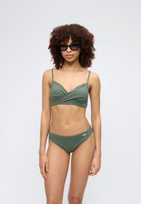 Γυναίκα με σγουρά μαλλιά, που φοράει μαύρα γυαλιά ηλίου και ελιές πράσινο μπικίνι με λεπτές τιράντες, στέκεται μπροστά σε απλό φόντο.