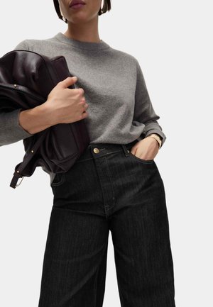 Suéter gris con un escote redondo, combinado con jeans de pierna ancha negros. Se sostiene una bolsa de cuero oscura en la mano izquierda, con un acento dorado.