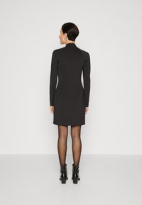 Robe noire à manches longues avec un col montant, au design ajusté et longueur au-dessus du genou, associée à des bottines noires élégantes et des collants transparents.