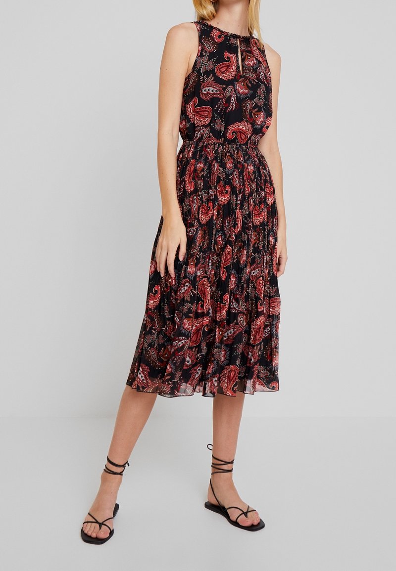 Femme portant une robe midi noire sans manches avec un motif paisley rouge et des sandales plates noires à brides, debout devant un fond gris.